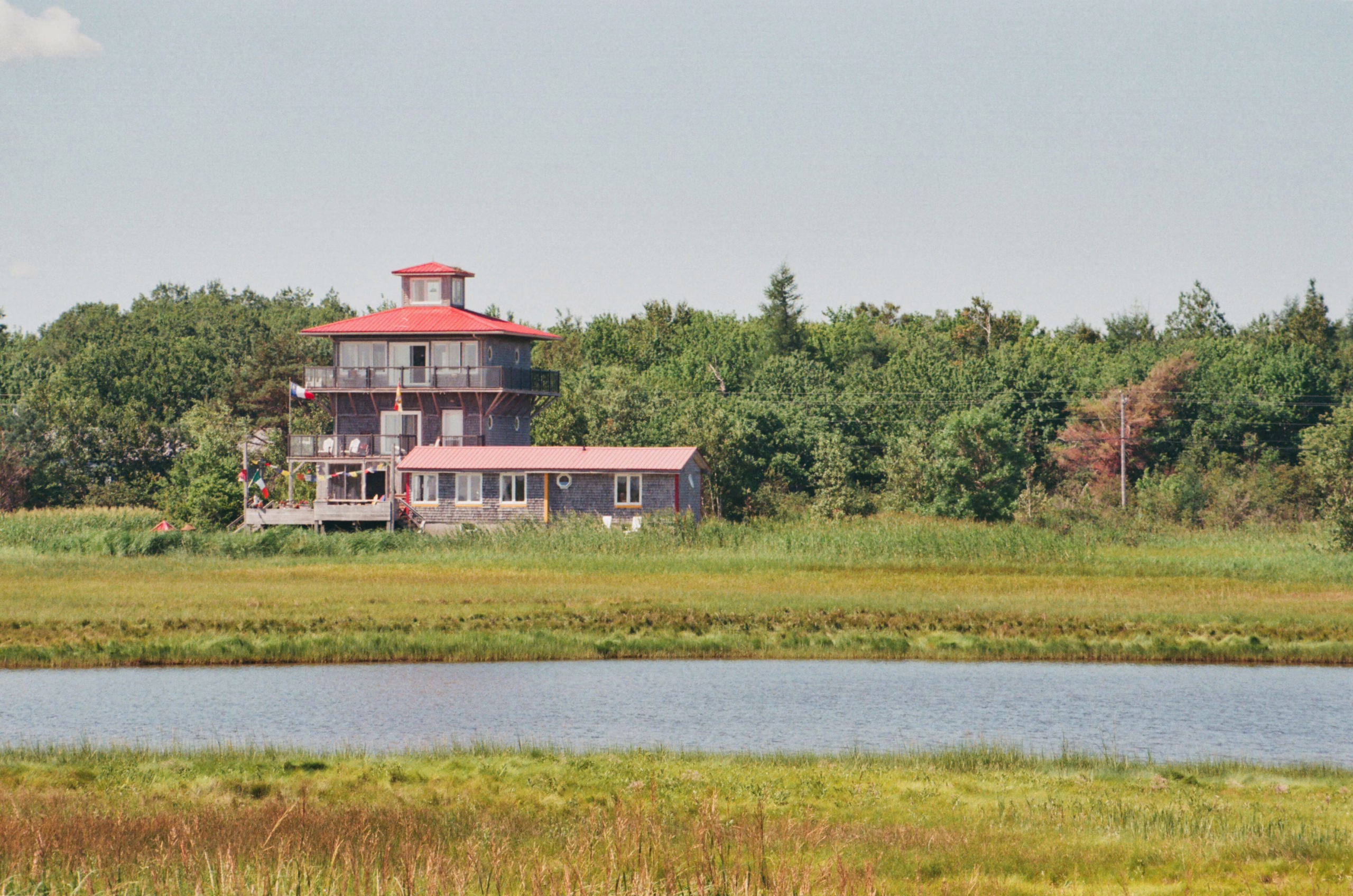 A multi-story gray shingled house with a distinctive red roof and cupola, situated between a dense forest and a tranquil marsh with water in the foreground.