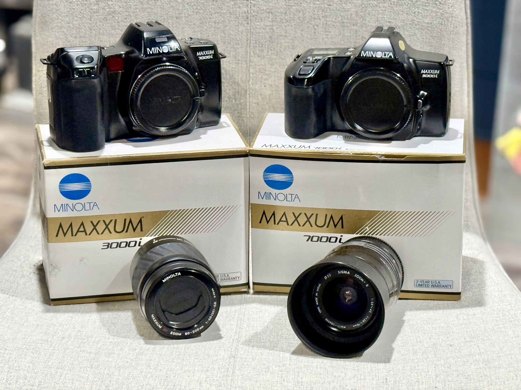 2 minolta cameras, a 3000i series and 7000i series both sitting on top of their respective boxes. In front of the boxes are 2 lenses, a 28-70mm and a 70-200mm respectively.